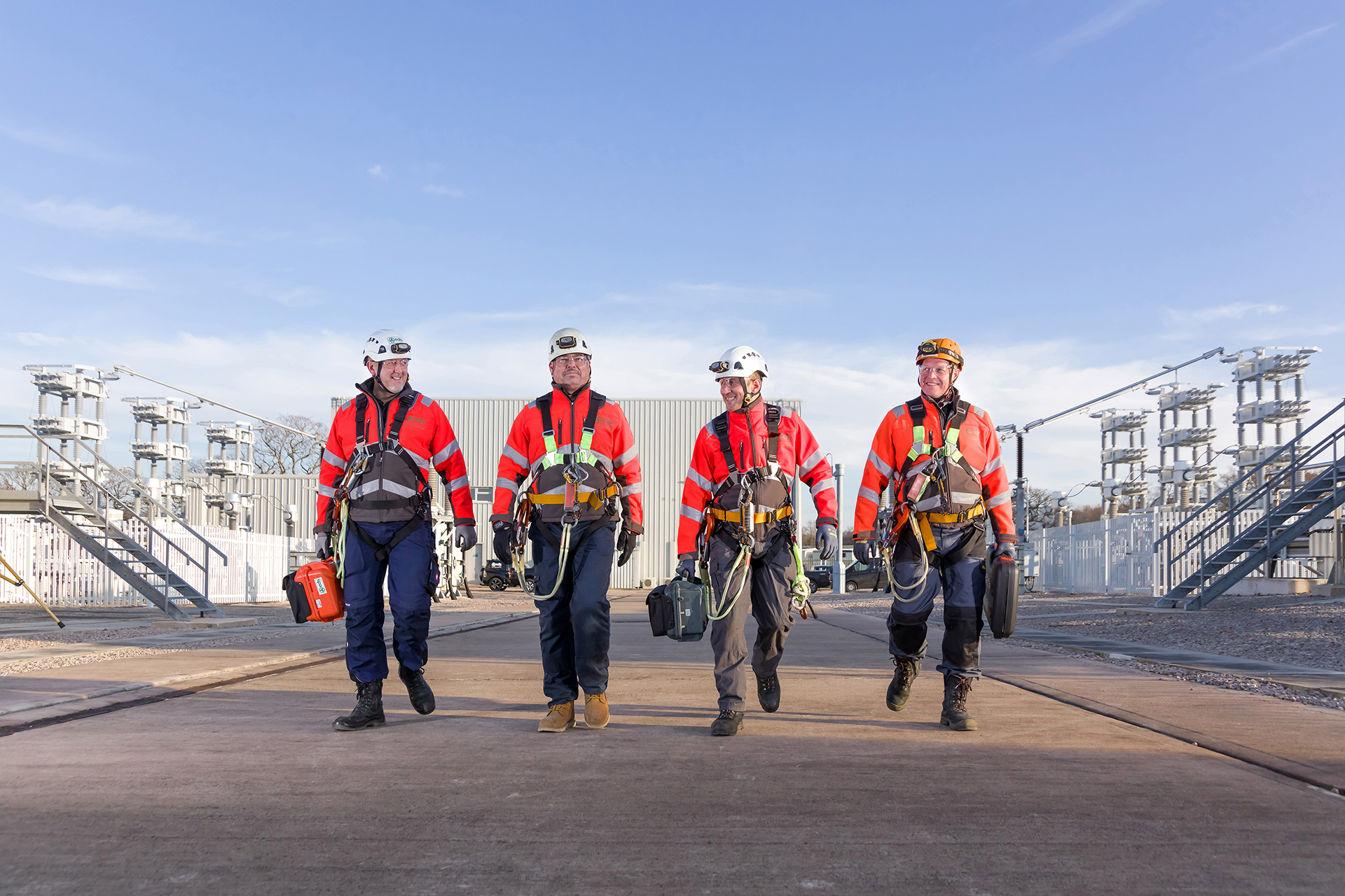 Four high voltage engineers walking in a line towards the camera with HV equipment in the background