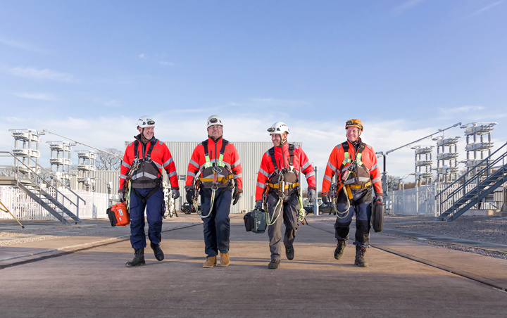 Four high voltage engineers walking in a line towards the camera with HV equipment in the background